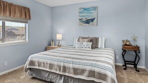 A bedroom with a bed, a nightstand, a lamp, a painting, and a window with blinds.