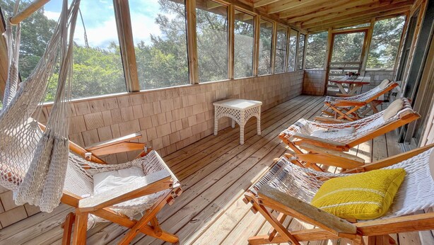 A wooden deck with hammocks and lounge chairs, a small table, and a view of trees through large windows. - Sunspace:  Quiet location, screened porch, deck, treetop sunsets (Ocracoke)