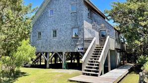 A rustic wooden house with a steep gable roof, a wooden deck, and a staircase leading to a covered porch. - Sunspace:  Quiet location, screened porch, deck, treetop sunsets (Ocracoke)