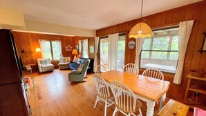 A cozy cabin interior with a wooden dining table, white chairs, a sofa, and a rocking chair. - Sunspace:  Quiet location, screened porch, deck, treetop sunsets (Ocracoke)