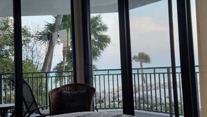 Uma varanda com cadeira de vime e uma mesa, com vista para uma costa rochosa com palmeiras.