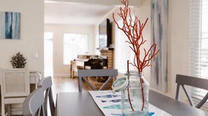 Una mesa de comedor con un tarro de cristal que contiene ramas, una silla blanca y otra mesa de comedor con un posavasos.