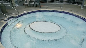 Een rond bubbelbad met betegelde rand, omgeven door een betonnen terras en metalen leuningwerk.