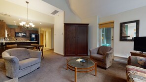 A living room with a round coffee table, two armchairs, a television, and a kitchen area with wooden cabinets and a microwave.