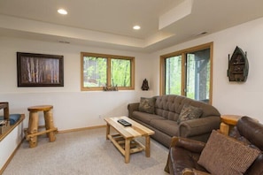 A cozy living room with a brown leather sofa, a wooden coffee table, and two wooden stools. There are two windows with wooden frames, one of which has a view of trees. A framed picture hangs on the wall, and a small wooden shelf is mounted on the wall.
