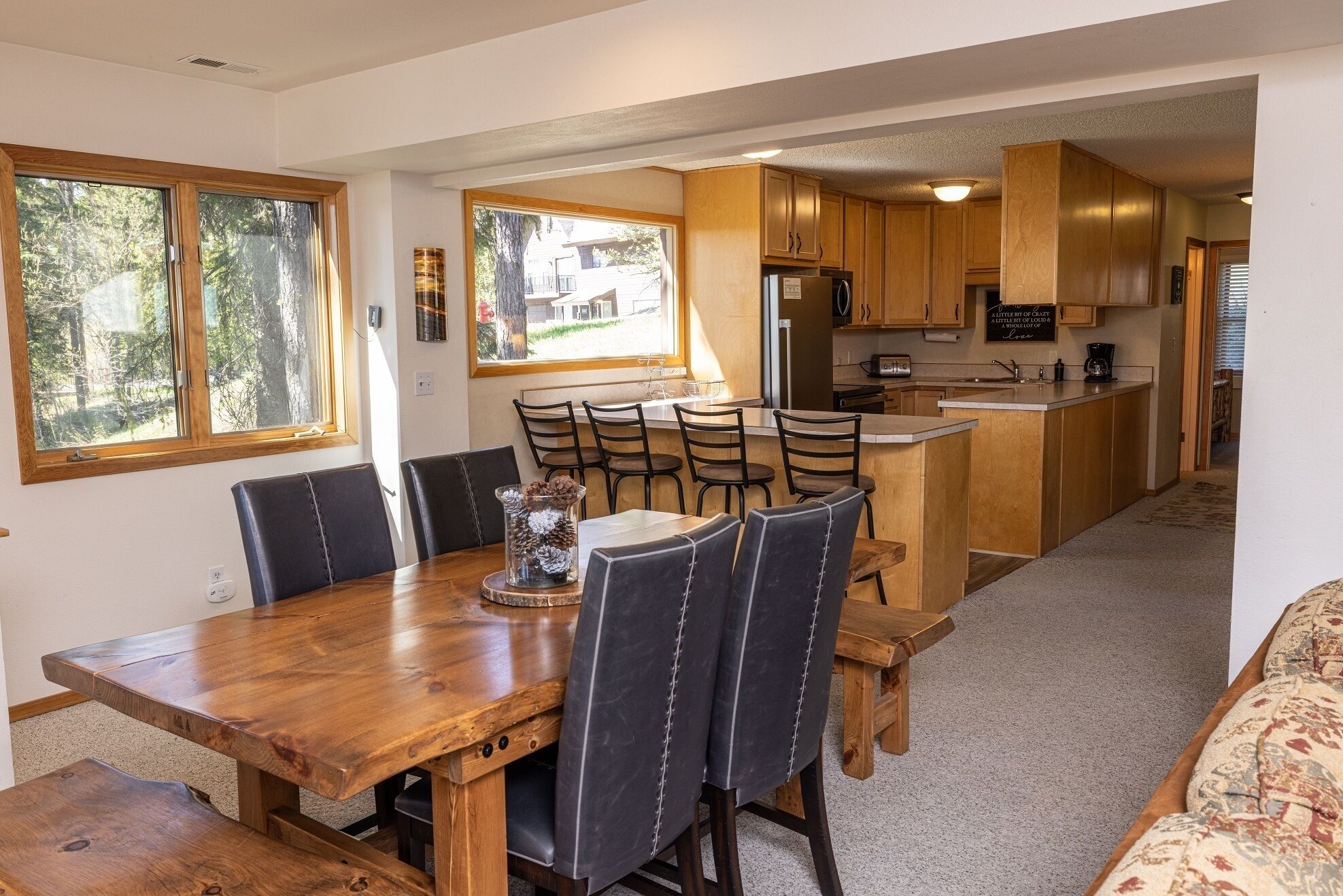 A dining area with a wooden table and chairs, a kitchen with wooden cabinets, and large windows offering a view of the outdoors.