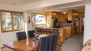 A dining area with a wooden table and chairs, a kitchen with wooden cabinets, and large windows offering a view of the outdoors. - Spacious Home with Ski Access (Whitefish)