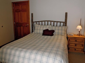 A bed with a plaid bedspread, a wooden headboard, a dark-colored pillow, and a bedside table with a lamp and a small electronic device.
