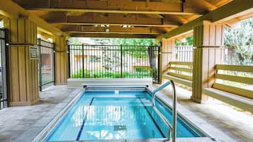 A covered swimming pool with a wooden deck, metal railings, and a view of a green garden.