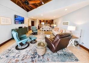 A spacious living area with a modern kitchen in the background, a television mounted on the wall, and a ceiling fan.