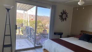 A bedroom with a bed, a ceiling fan, a nightstand, a window with a view of mountains, and a patio area with a chair and table. - Lake view home with mountain views by Havasu Springs (Parker)