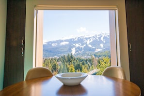 A dining table with a bowl in the center, a window with a view of snow-capped mountains and forested valley, and two chairs.