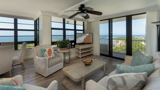 A spacious living room with a large TV, comfortable seating, and a balcony with a view of the ocean.
