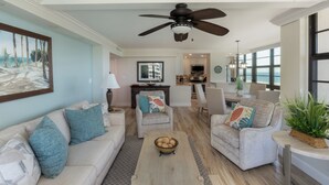 A spacious living room with a ceiling fan, light-colored furniture, and a view of the kitchen and dining area.