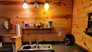 A wooden cabin kitchen with a sink, countertop, and wooden cabinets. There are overhead lights, a clock, and a shelf with various items. - Black Hills Get-Away! (Custer)