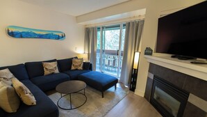 A living room with a sectional sofa, a round coffee table, a fireplace, and a balcony with curtains. - Renovated, Whistler Village Condo, Private Hot Tub, BBQ & Pool Access. (Whistler)