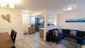 A modern living room with a dark blue sofa and a coffee table, a wooden dining table with chairs, and a kitchen in the background. - Renovated, Whistler Village Condo, Private Hot Tub, BBQ & Pool Access. (Whistler)