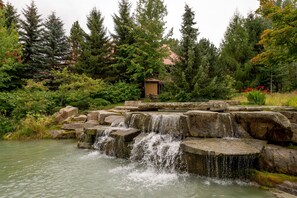 A natural water feature with cascading water over rocks, surrounded by dense greenery and trees. - Renovated, Whistler Village Condo, Private Hot Tub, BBQ & Pool Access. (Whistler)