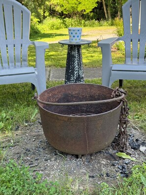 A rustic outdoor setting with a large metal pot, two white chairs, and a patterned table.