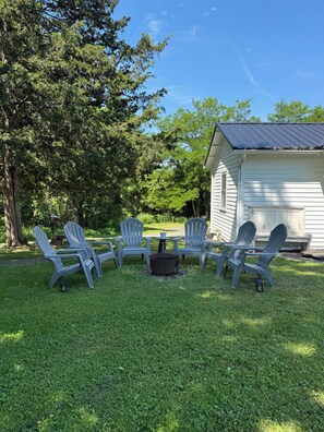A grassy outdoor area with Adirondack chairs arranged around a fire pit, a small table, and a white building with a dark roof.