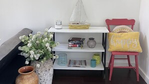A living room with a wooden floor, a sofa, a red chair, a white shelf with books and decorative items, a vase with flowers, and three framed pictures on the wall.