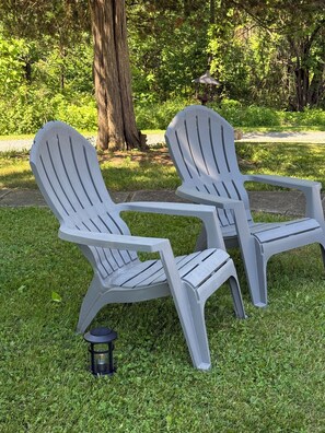 Two Adirondack chairs with a small table between them, set on a grassy area with a tree and a lantern in the background.