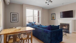 A living room with a blue sectional sofa, a fireplace, a dining table with chairs, and a TV mounted above the fireplace. - The Upscale Getaway Near Downtown (Chicago)