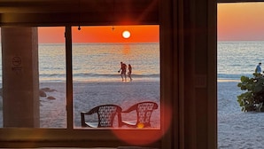 A room with a large window overlooking a beach at sunset, featuring two chairs and a bed.