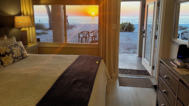 A bedroom with a bed, a nightstand, a lamp, and a view of the beach at sunset.