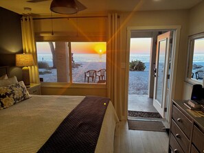 Un dormitorio con cama, mesita de noche, lámpara y vista al mar al atardecer.