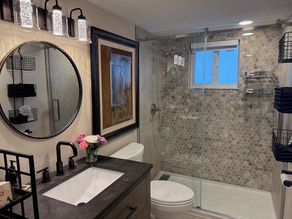 Un baño con un espejo redondo, un lavamanos blanco, un inodoro y una zona de ducha con mampara de vidrio y pared de mosaico de piedra.