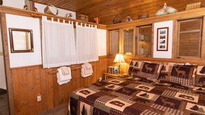 Ein Zimmer mit Holzwänden, einem Bett, einem Fenster mit Jalousien, einer Lampe und einer gesteppten Bettdecke.