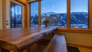 Un tavolo da pranzo in legno con panche, un vaso con fiori e vista su montagne innevate attraverso ampie finestre.