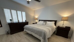 Un dormitorio con una cama grande, dos mesitas de noche, un aparador, una ventana con persianas y un ventilador de techo.