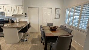Cocina moderna con armarios blancos, encimera de granito y horno empotrado. Cuenta con una zona de comedor con mesa de madera, sillas tapizadas en gris y un cuenco decorativo.