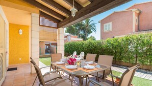 Un patio cubierto con una mesa lista para comer, rodeado de sillas y vistas a una zona residencial con una casa y vegetación.