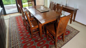 A dining area with a wooden table and chairs, a patterned rug, and a view of greenery through large windows.