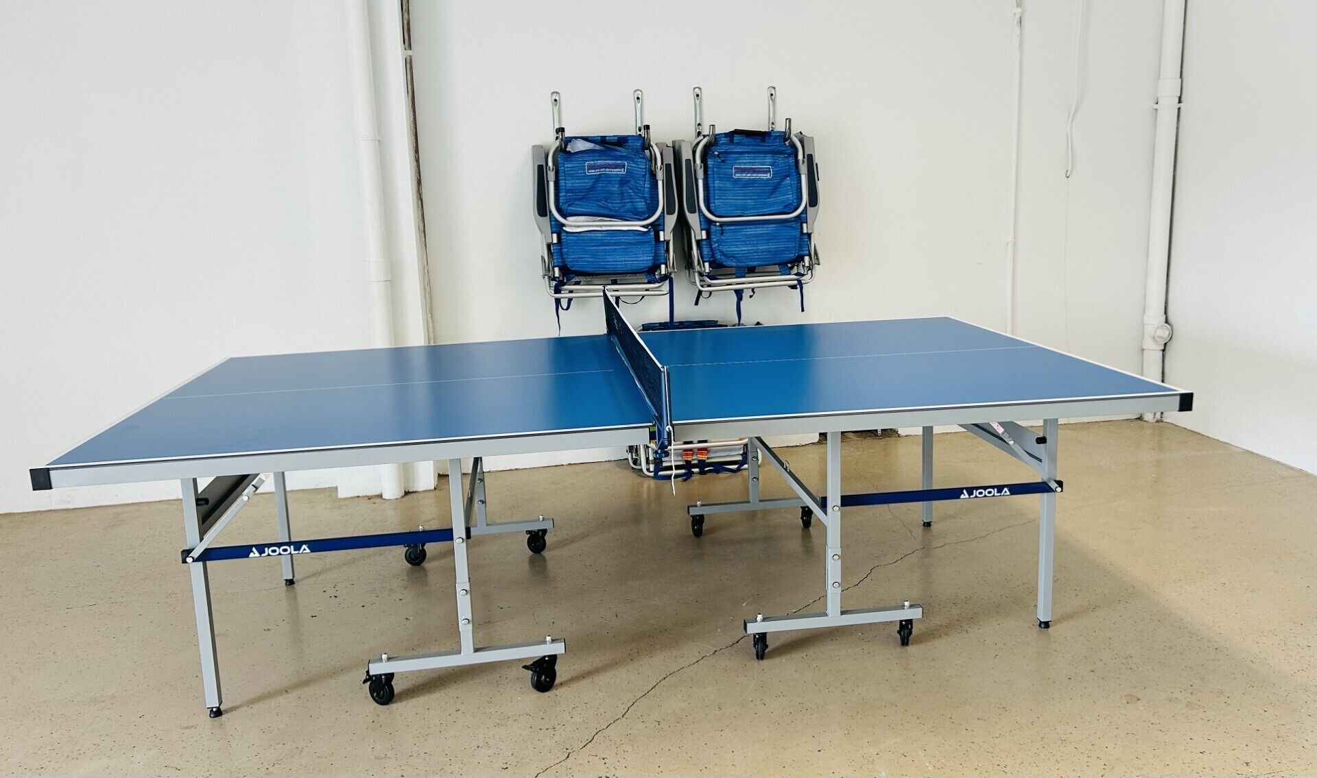 A portable ping pong table with wheels, two blue chairs, and a white wall in the background.