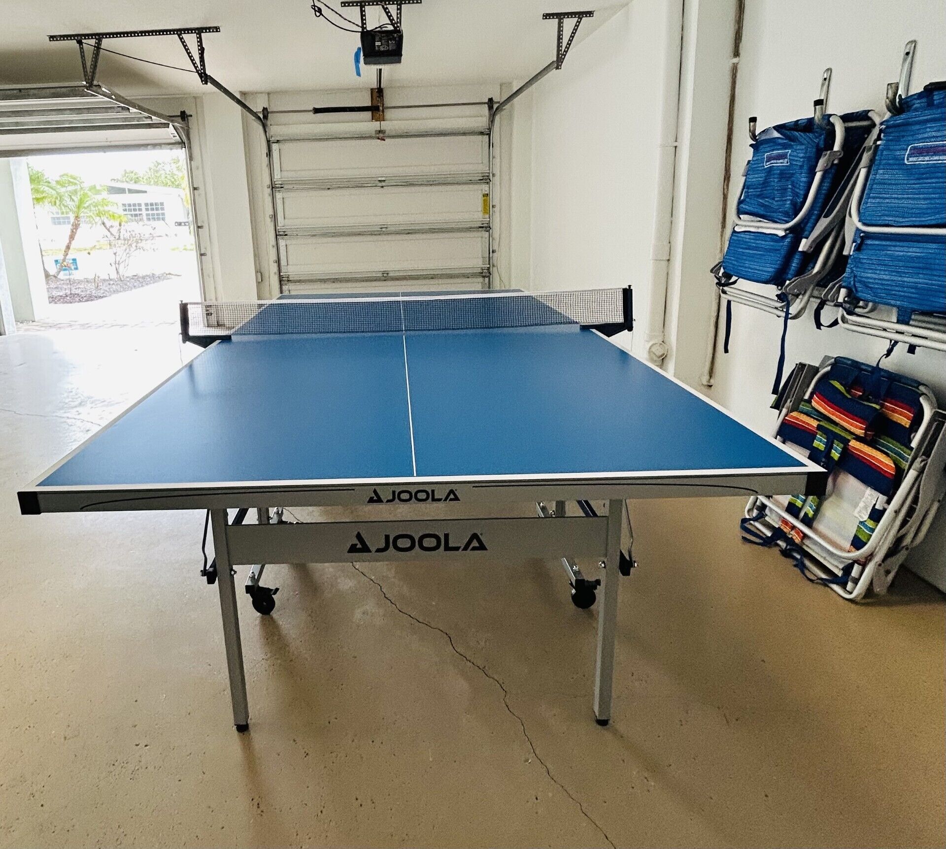 A ping pong table with a white frame and blue playing surface, labeled 'AJOOLA'.