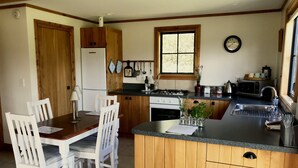 A kitchen with wooden cabinets, a dining table with chairs, a refrigerator, a stove, and a window with a clock.