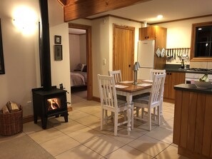 A cozy living room with a fireplace, a dining table with chairs, a kitchen area with wooden cabinets, and a bed in the background. - Dara Vineyard Cottage Martinborough (Martinborough)