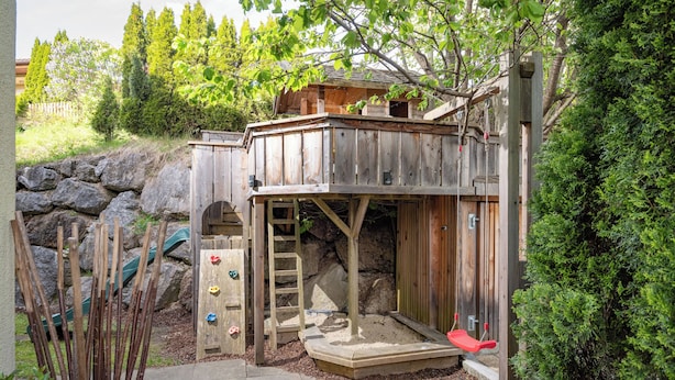 Une cabane en bois, dotée d’un toboggan et d’un mur d’escalade, entourée de rochers et de verdure.