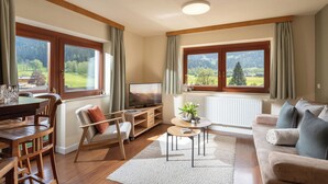 Un salon comprenant une table à manger en bois, un canapé, un téléviseur et de grandes fenêtres donnant sur un paysage verdoyant.