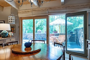 Un coin repas comprenant une table ronde en bois, des chaises, une lampe décorative et une vue sur un jardin grâce à de grandes portes vitrées.