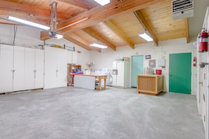 Een ruime kamer met hoge plafonds, houten balken en een betonnen vloer. Er is een kitchenette met een koelkast, een wastafel en een houten tafel. Op de achtergrond is een groene deur en een witte kast zichtbaar.