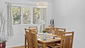 Un comedor con una mesa de vidrio, sillas de mimbre, una lámpara colgante y vistas a árboles por las ventanas.