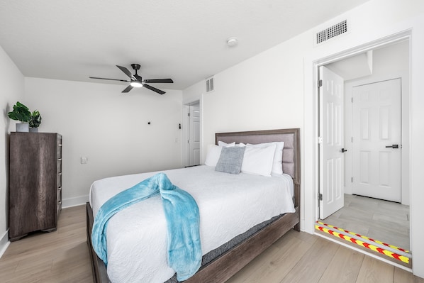 A bedroom with a bed, a ceiling fan, a door, and a small plant on a cabinet. - Bright Beach Cottage w/ Patio l Steps to the Beach & Restaurants l Pet-Friendly (Clearwater)