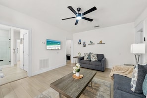 A modern living room with a flat-screen TV, a grey sofa, a coffee table, and a ceiling fan. - Bright Beach Cottage w/ Patio l Steps to the Beach & Restaurants l Pet-Friendly (Clearwater)