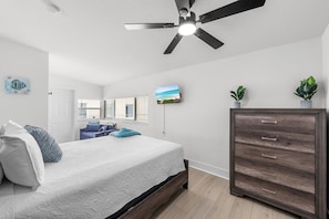 Un dormitorio con cama, ventilador de techo, televisor, cómoda y ventana con persianas.