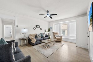 A modern living room with a sofa, coffee table, and armchairs, a ceiling fan, and large windows with blinds.
